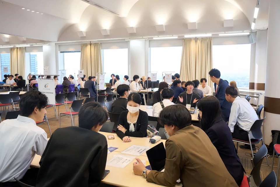 企業名のパネルが並ぶ会場で、複数のテーブルを数人ずつで囲んで話す参加者